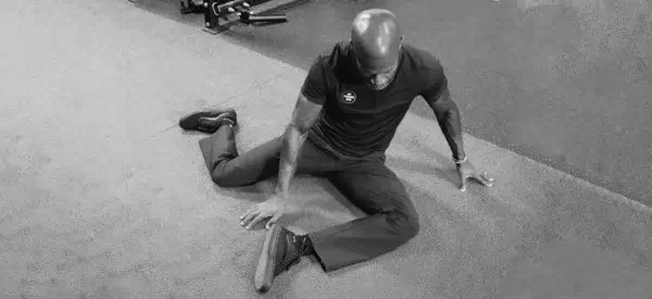 A person is seated on a carpeted floor, performing the 90-90 stretch to improve hip mobility, with one leg bent in front and the other behind, using their arms for support.