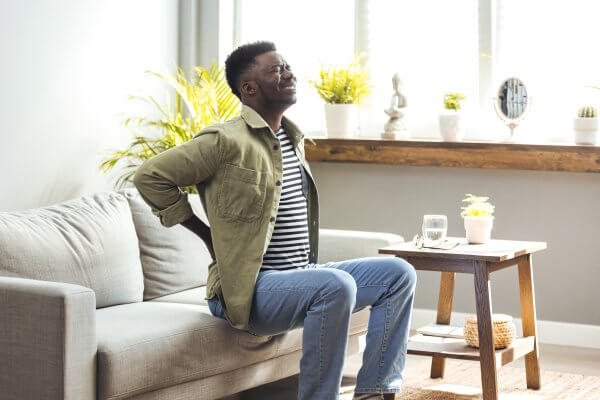 Man sitting on a couch at home with hand on his lower back, expressing discomfort and in need of Couch Cure.
