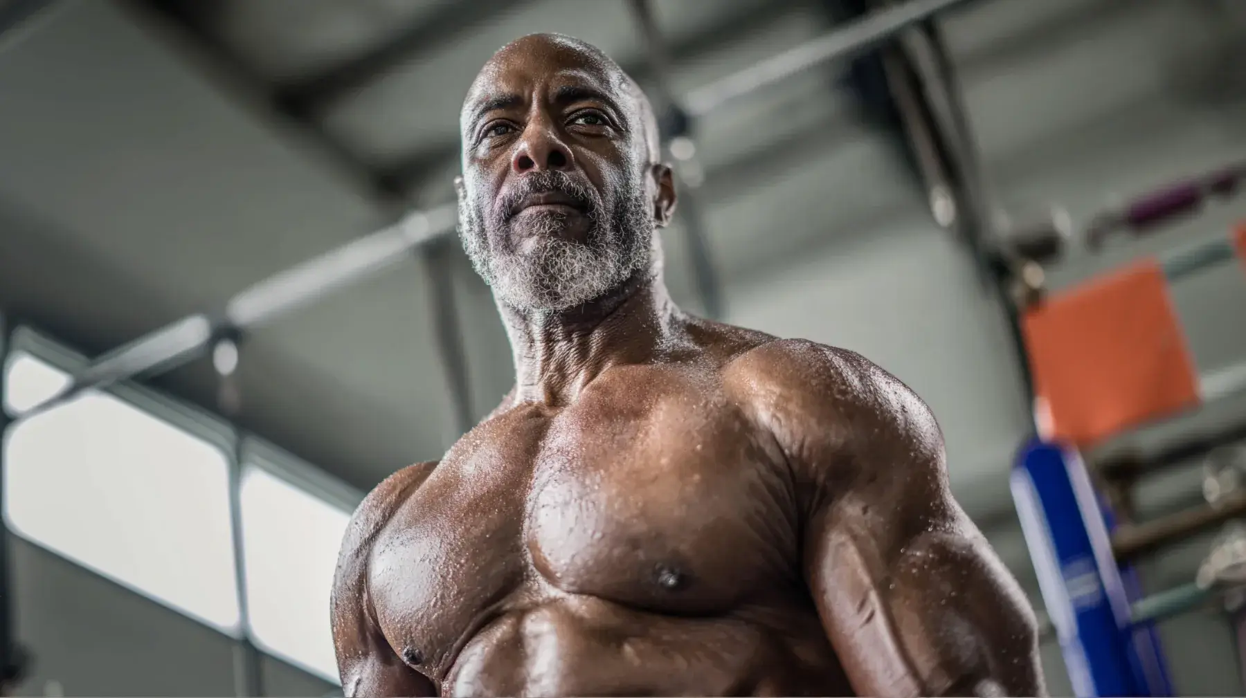 A muscular, middle-aged man with a gray beard stands shirtless in a gym, his skin glistening with sweat and workout equipment visible in the background.