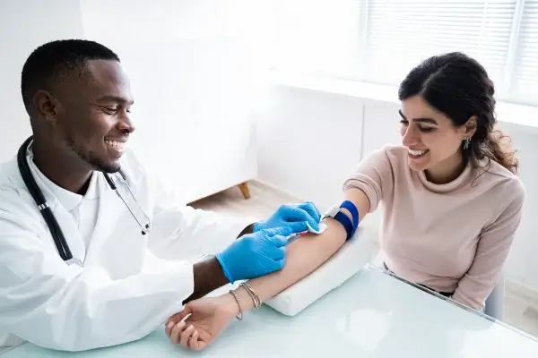 A healthcare professional prepares to draw blood from a smiling woman who is seated, with a tourniquet on her arm, in a bright medical office.