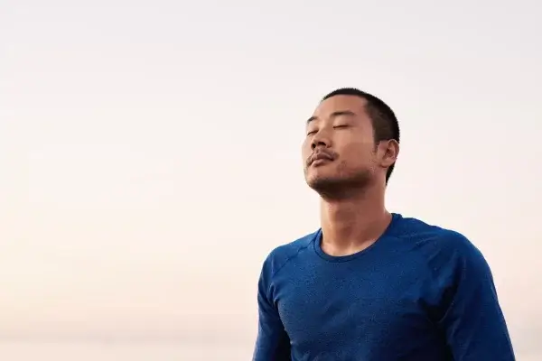 A man in a blue shirt meditates with closed eyes, a calm expression, and stress-reducing breathing techniques under a soft pink sky in the background.