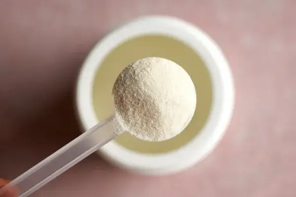 A close-up of a white plastic scoop filled with a fine, white collagen powder, held above an open container with a similar substance inside. The background is a soft, out-of-focus pink surface.