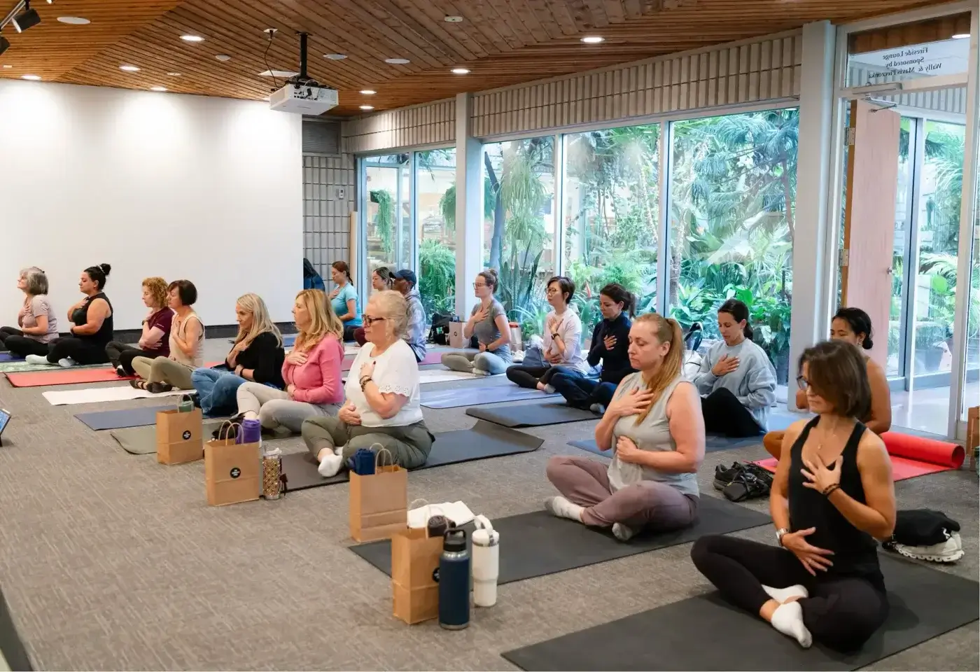 A group of people sit cross-legged on yoga mats in a spacious room, each with one hand on their chest and the other on their abdomen, practicing mindful breathing.