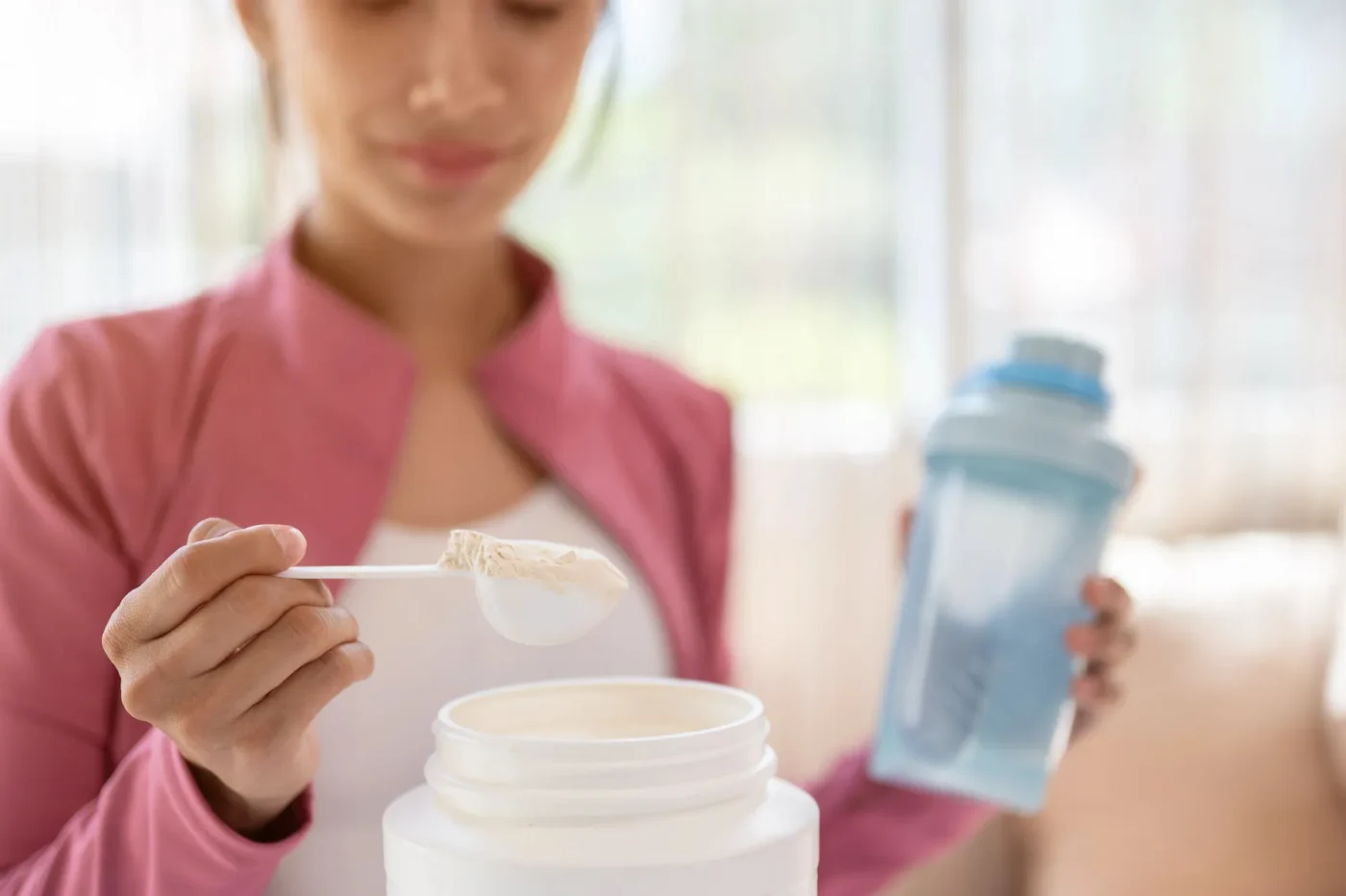 A woman in a pink jacket holds a shaker bottle in one hand and scoops protein powder from a container with the other hand, preparing a drink.