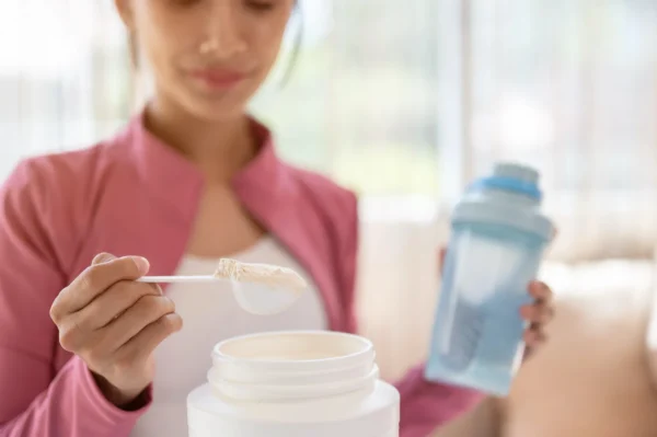 A woman in a pink jacket holds a shaker bottle in one hand and scoops protein powder from a container with the other hand, preparing a drink.