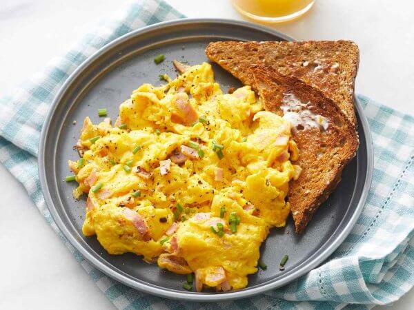Plate of scrambled eggs with chives and bits of ham, served with a slice of buttered whole-wheat toast on a checkered cloth. This delicious breakfast is both flavorful and mindful of health, offering a satisfying start to your day.