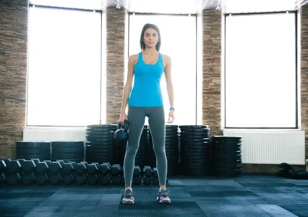A woman holding dumbbells stands in a gym with large windows and a brick wall, wearing a blue tank top and gray leggings, breathing deeply as she exercises.
