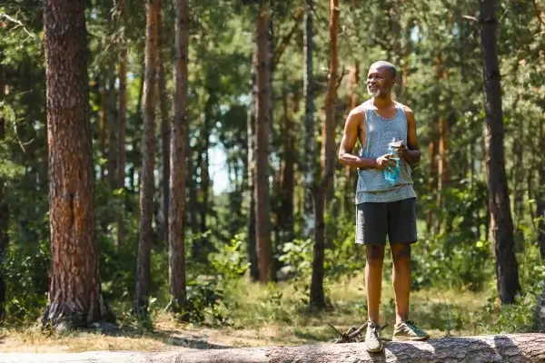 A man stands on a log holding a water bottle in a forest, wearing a tank top and shorts, with trees in the background. The serene surroundings remind him of the healing power of music that often accompanies his woodland hikes.