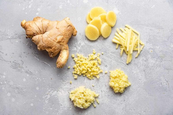 Fresh ginger root, an ancient spice hailed as a modern health miracle, next to its different preparations: sliced, julienned, minced, and grated on a grey surface.