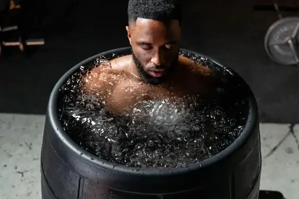 A man is submerged up to his shoulders in a barrel filled with ice water, embracing cold therapy. He appears to be exhaling deeply, with fitness equipment visible in the background.
