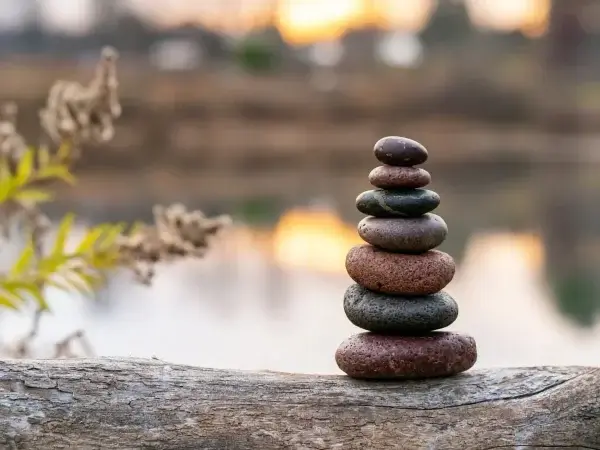 A stack of seven smooth stones balanced on a log near a calm body of water, symbolizing control and acceptance, with blurred greenery and sunset in the background.
