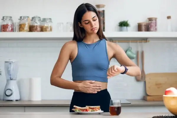 A woman in a kitchen checks her watch while placing a hand on her stomach, fasting as a sandwich and a glass of juice await on the table.