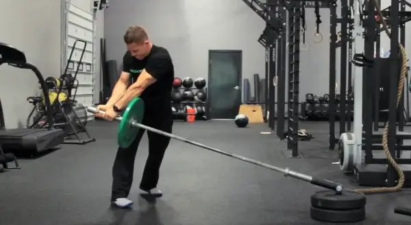 A person stands in a gym performing a Landmine Twist with a barbell anchored at one end and a green weight plate on the other, in a spacious workout area with various fitness equipment to build core strength.