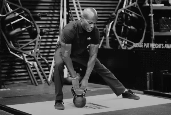 A man in a gym performing a lateral lunge with a kettlebell, focusing intently on his form, dressed in athletic gear. The environment is equipped with various workout machines and neatly arranged weights.
