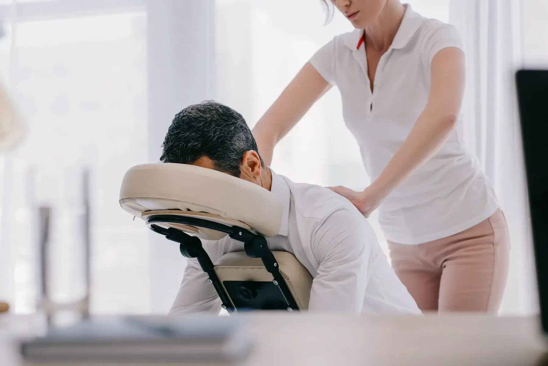 A person receives a shoulder massage while seated in a portable massage chair, administered by another person in a light-colored top and pants in a bright room.