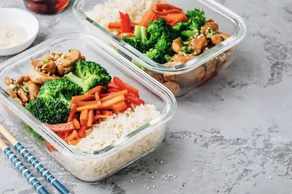 Two glass meal prep containers filled with white rice, broccoli, carrots, red bell peppers, and chicken teriyaki are on a gray surface, with chopsticks and a bowl of sesame seeds nearby. These vibrant dishes are perfect for healthy eating and provide great meal prep ideas.