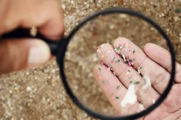 A hand holding microplastics is viewed through a magnifying glass, showcasing small colorful plastic particles against a background of sand, underscoring the environmental impact of these tiny pollutants.