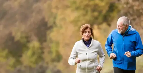 An older man and woman jogging together in a park, wearing sportswear, with a blurred green background, demonstrating their commitment to longevity fitness.