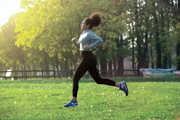 A person runs through a grassy park with trees in the background, wearing athletic clothing and sneakers, bathed in sunlight, enjoying the benefits of nature's gym.
