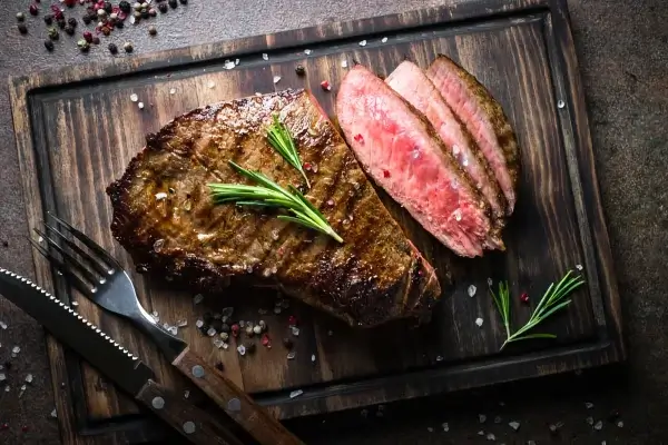 A sliced, medium-rare steak garnished with rosemary on a wooden cutting board, accompanied by a fork and knife. Perfect for those mindful of carbs, this red meat dish is both savory and satisfying.