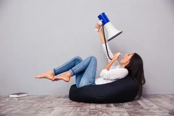 A woman reclining on a black bean bag and playfully shouting into a blue megaphone, exploring self-talk techniques, with a smartphone beside her on the floor.