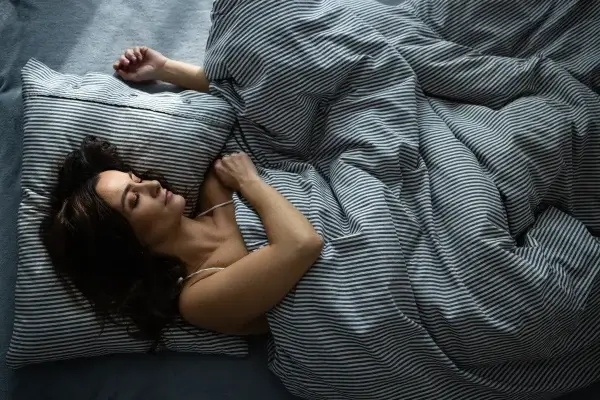 A person is lying in bed, covered with a striped comforter and resting on a matching pillow. They appear to be asleep, basking in the restful benefits that contribute to their longevity.
