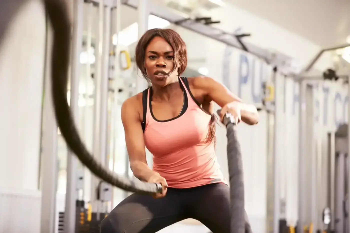 A woman wearing a pink tank top and black leggings exercises with battle ropes in a gym.