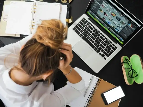 Person with a bun hairstyle looks stressed while working at a desk with a laptop displaying digital images, surrounded by notebooks, a phone, and eyeglasses.