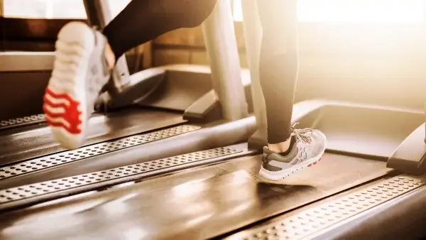Close-up of a person running on a treadmill at a gym, with sunlight streaming in. The person, focused on longevity, is wearing athletic shoes and leggings as they push through longer sprint intervals.