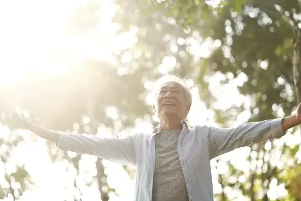 An elderly person stands outdoors with arms outstretched, smiling and facing the sunlight to soak in Vitamin D, surrounded by trees.