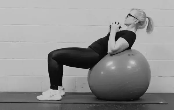 A person wearing glasses and athletic clothing is performing weighted crunches by lying back on a stability ball, with their feet flat on the ground and hands clasped together at their chest.
