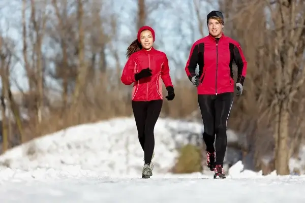 Two people jog outside on a snowy path, wearing red jackets, black pants, and winter accessories. Leafless trees are in the background.